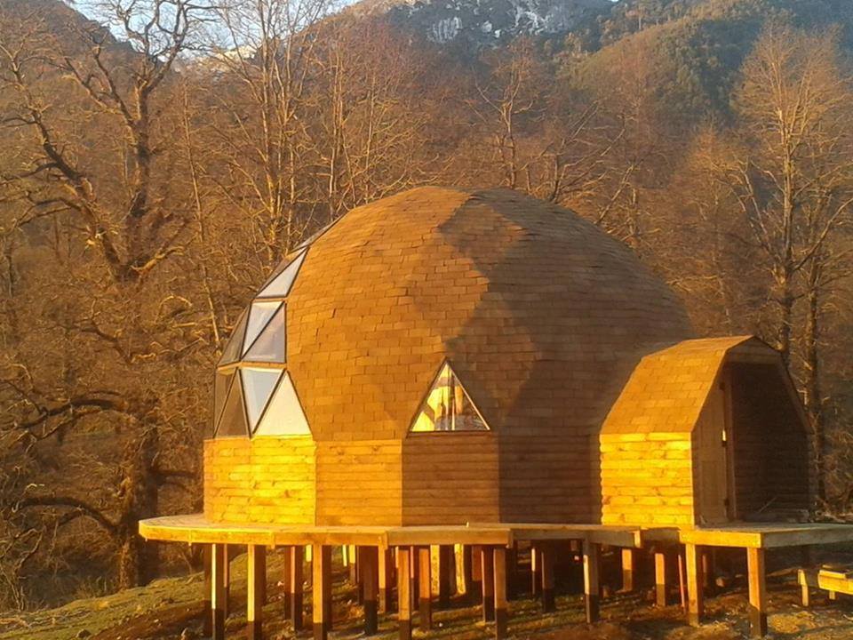 Domos Malalcahuello: domo geodésico de madera sobre una terraza elevada, entre árboles y cerros al atardecer