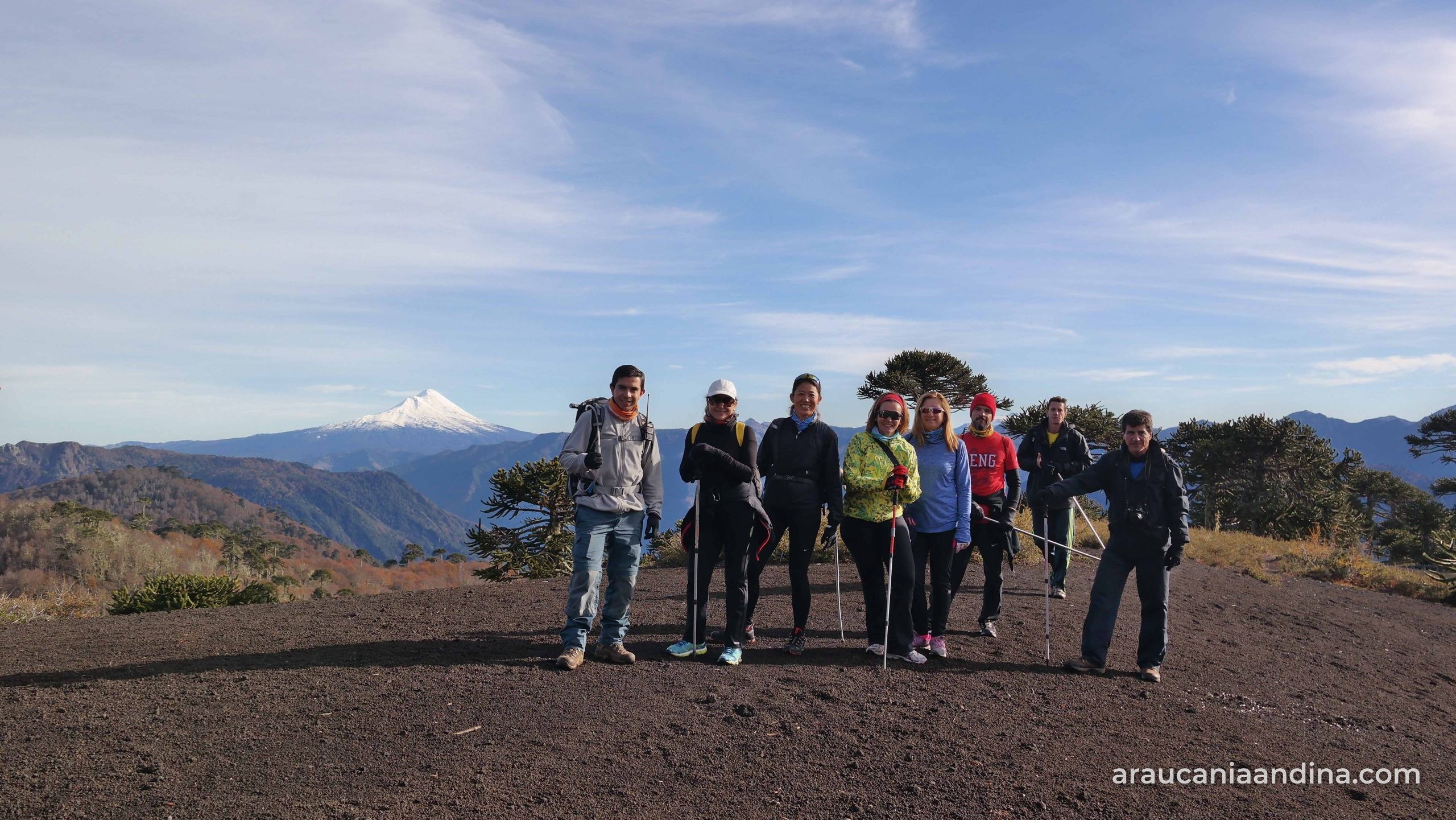 Grupo de excursionistas de Turismo Pewen Mapu sobre suelo volcánico entre araucarias, con un volcán nevado al fondo